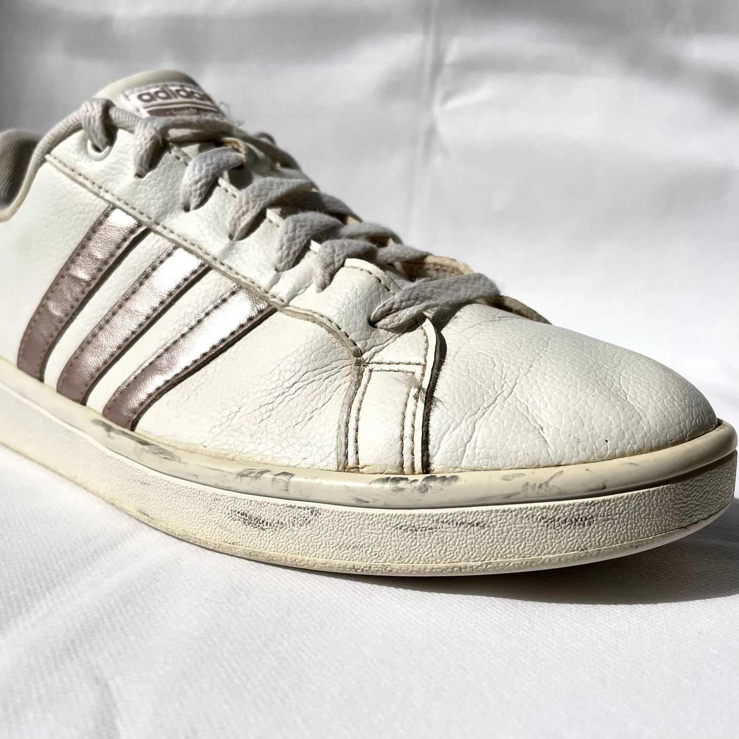 A close-up of a vintage Adidas sneaker in off-white leather with brown stripes, showing signs of wear and scuffing on the sole and toe, set against a wrinkled white fabric background.