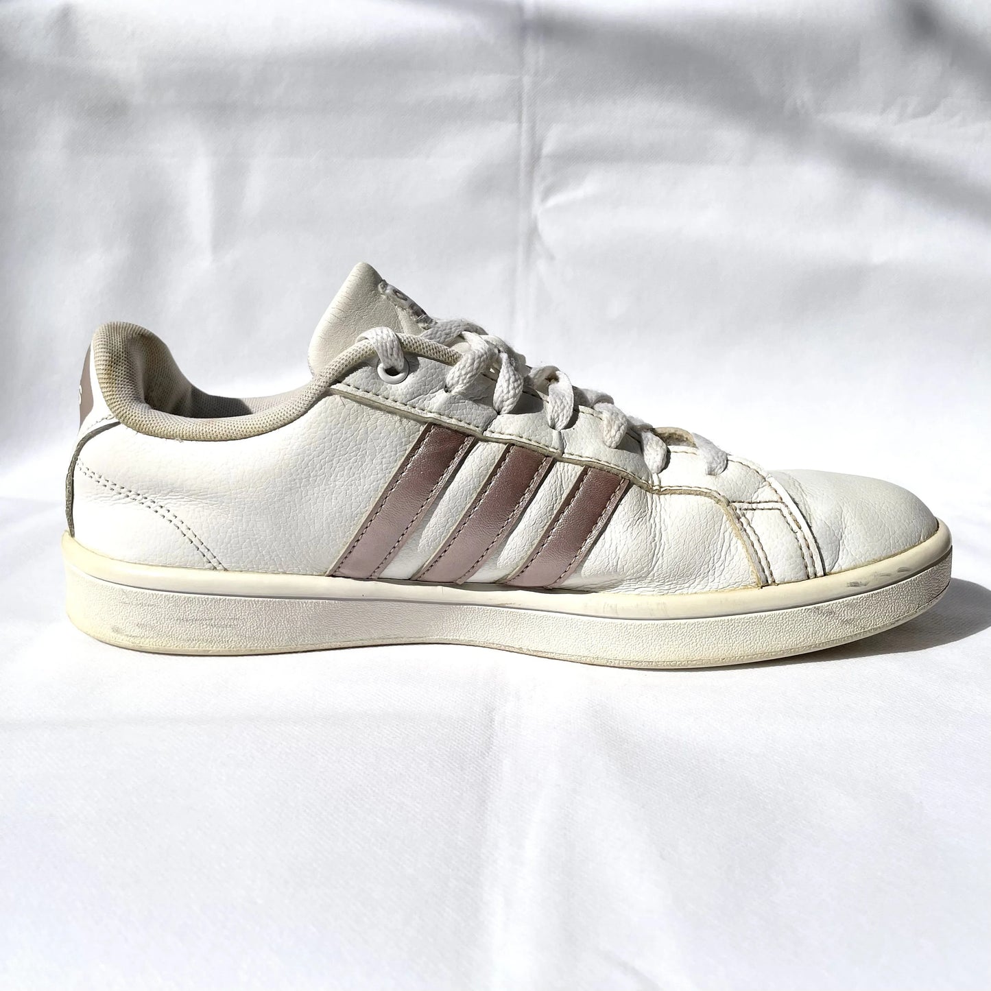 A single white leather sneaker with three metallic rose gold stripes on the side, photographed from the side profile against a wrinkled white fabric background.