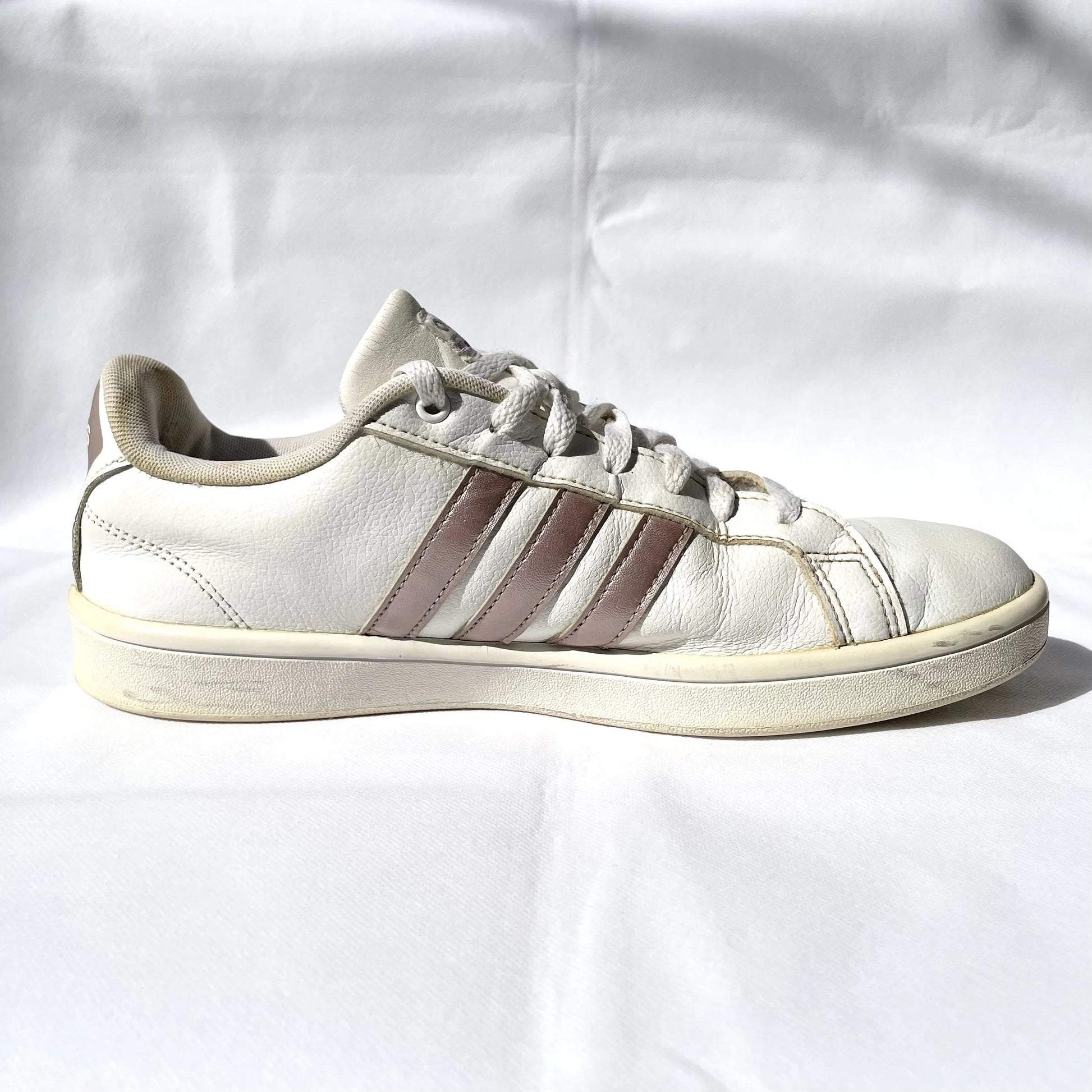 A single white leather sneaker with three metallic rose gold stripes on the side, photographed from the side profile against a wrinkled white fabric background.