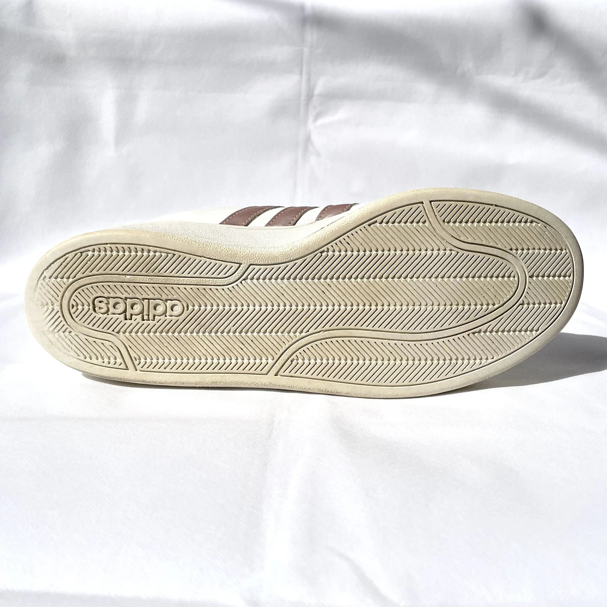The outsole of a vintage Adidas slide shoe, featuring the embossed brand name 'GODIPO' and three classic brown stripes on a cream-colored shoe, set against a wrinkled white fabric background.
