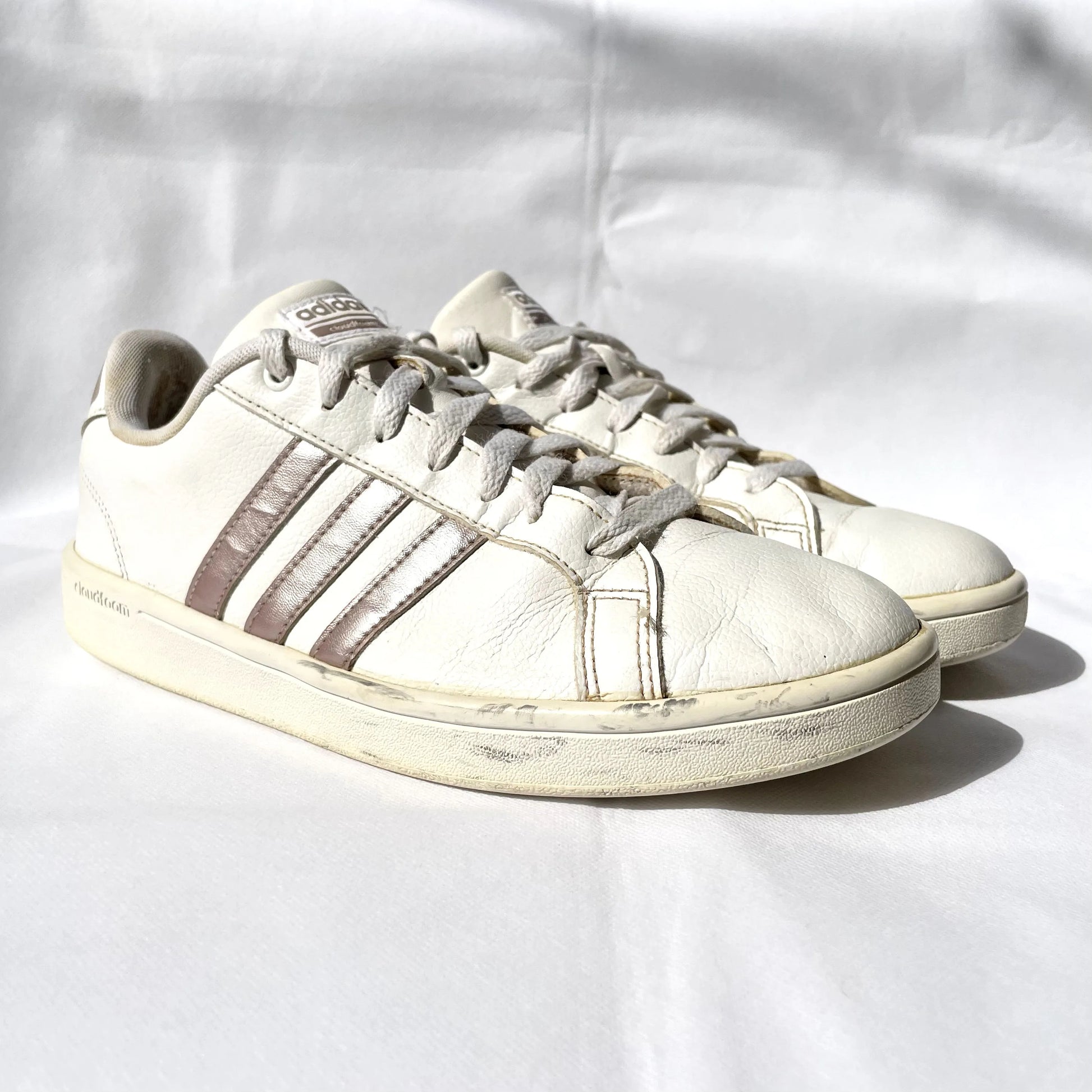 A pair of vintage Adidas low-top sneakers in off-white leather with brown stripes, displayed on a wrinkled white fabric background.