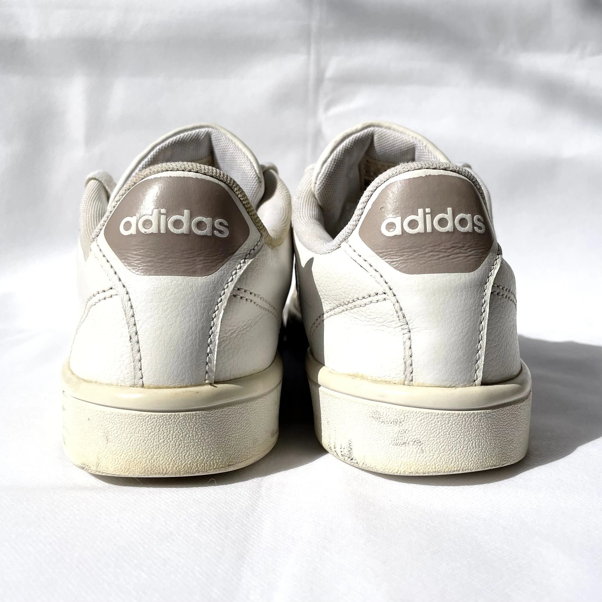 A pair of white Adidas sneakers viewed from the back, showing the heel area with white 'adidas' logos on taupe heel tabs and off-white soles, set against a white fabric background.
