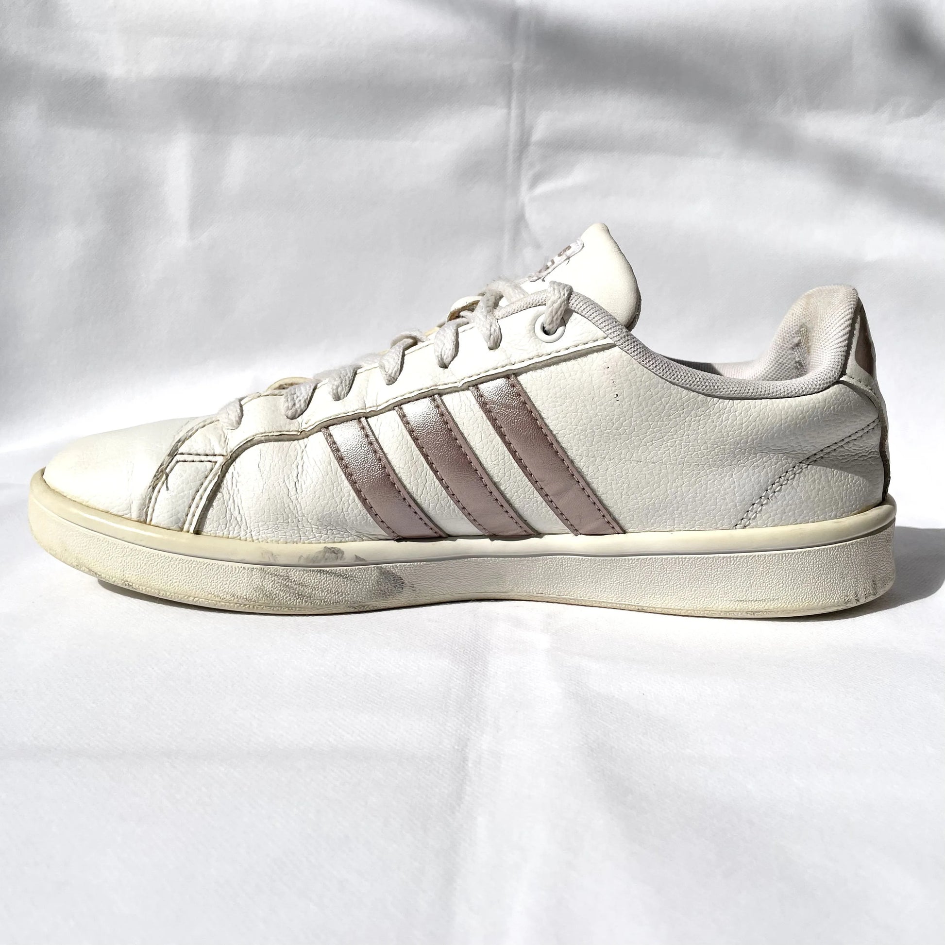 A single Adidas low-top sneaker in off-white leather with three metallic silver stripes, shown in side profile against a wrinkled white fabric background.