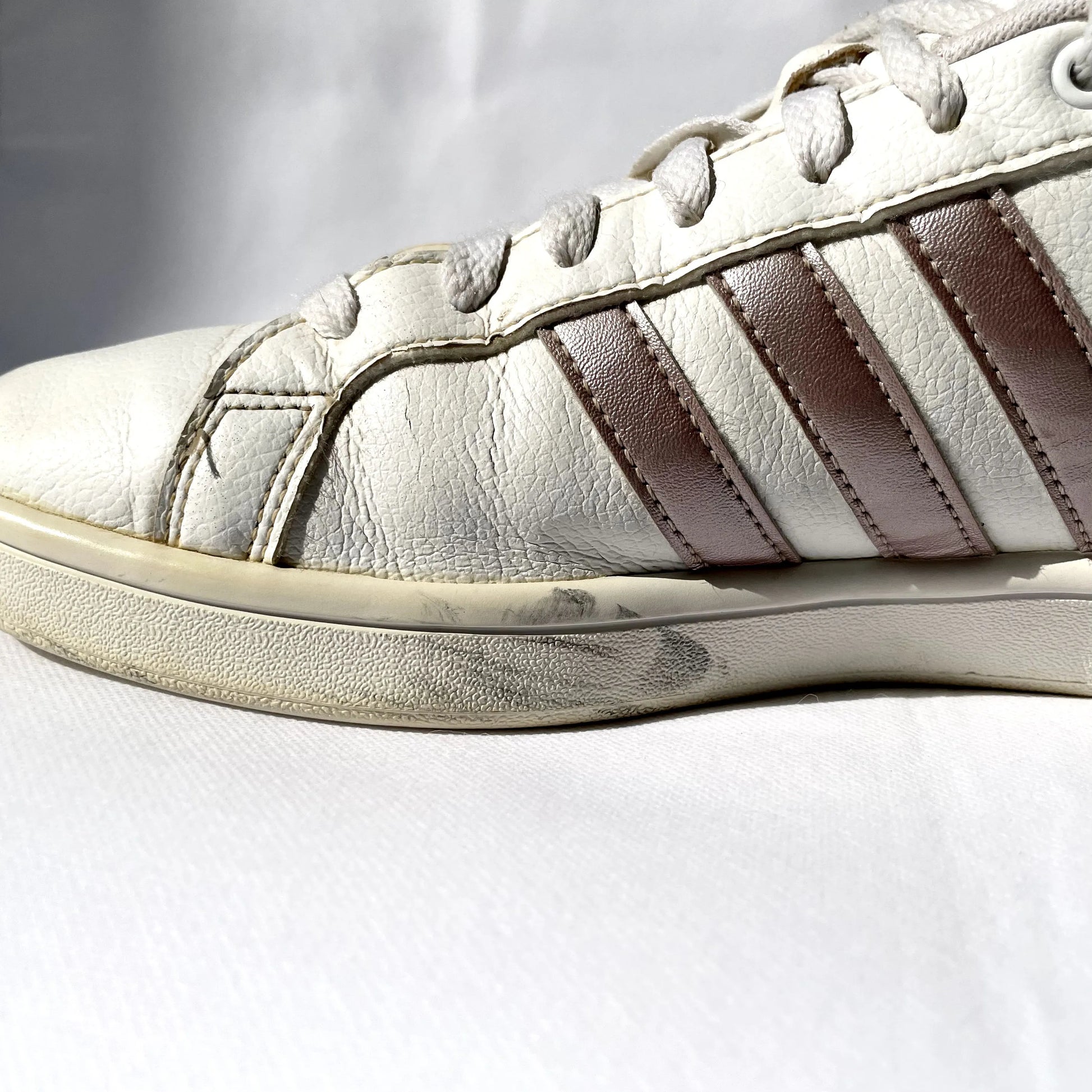 A close-up of a white leather sneaker with three dark brown stripes on the side, white laces, and a white rubber sole, photographed against a white fabric background.